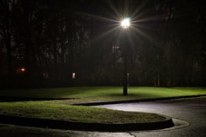 Lamp post at night, casting light on a wet, empty parking lot & lawn